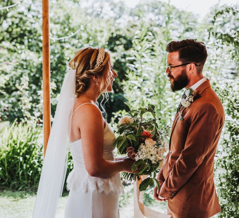 Bride & groom look lovingly at one another in front of green foliage background under tipi