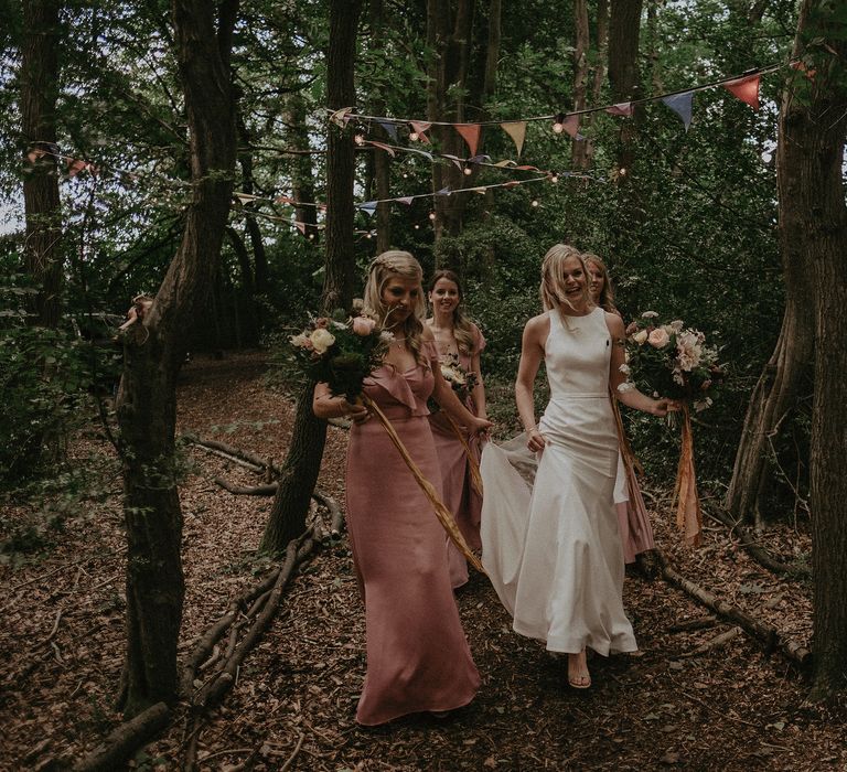 Bride laughs and smiles as she walks through woodland surrounded by her bridesmaids who wear pink gowns and hold floral bouquets