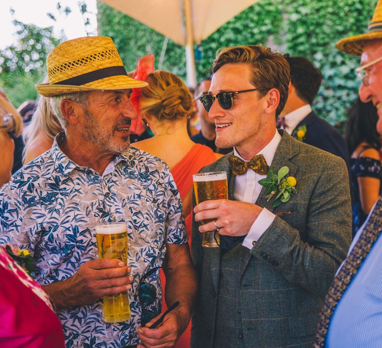 Grooms speaks to wedding guest as he holds pint of beer and wears sunglasses  | Story + Colour