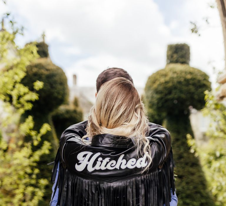 Groom in a blue suit embracing his bride at Euridge Manor in a fringed leather jacket with Hitched sign sticked on the back 