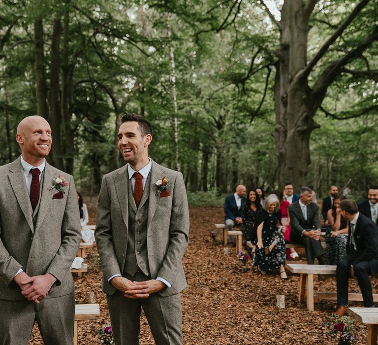 Groom and groomsmen in grey herringbone three piece suits, red ties and pocket squares and floral buttonholes wait for bride at woodland wedding ceremony in Norfolk