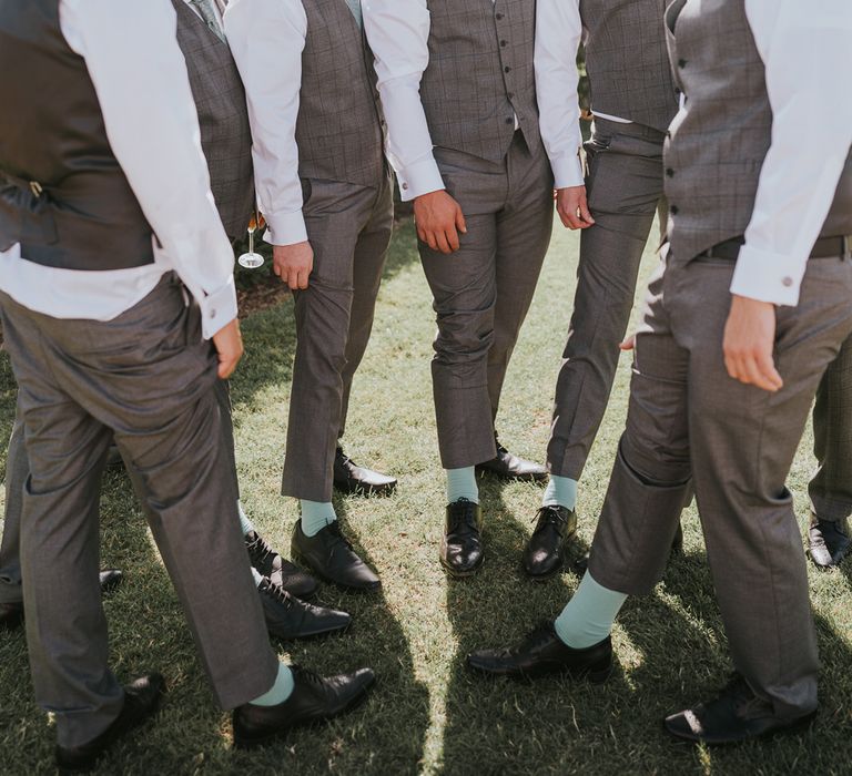 Grooms in grey waistcoats and trousers all pull them up to show mint green socks during summer wedding at Primrose Hill Farm