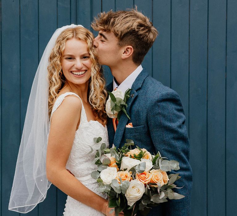 Bride looks toward camera as groom kisses her cheek on their wedding day