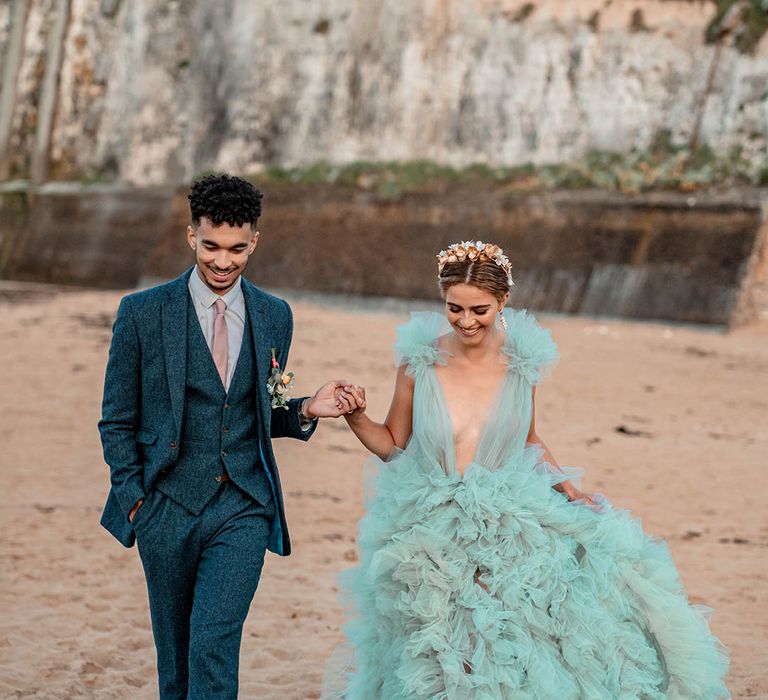 Groom in a blue wool suit holding hands with his bride in a layered tulle mint green wedding dress at their beach elopement 