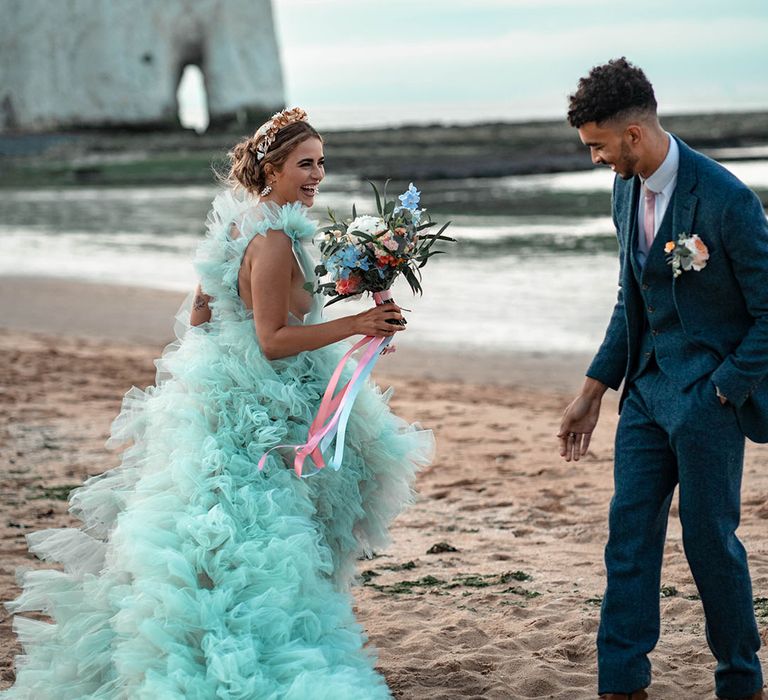 Groom in a blue wool suit smiling at his bride in a layered tulle mint green wedding dress as she dances on the sand at their coastal wedding 