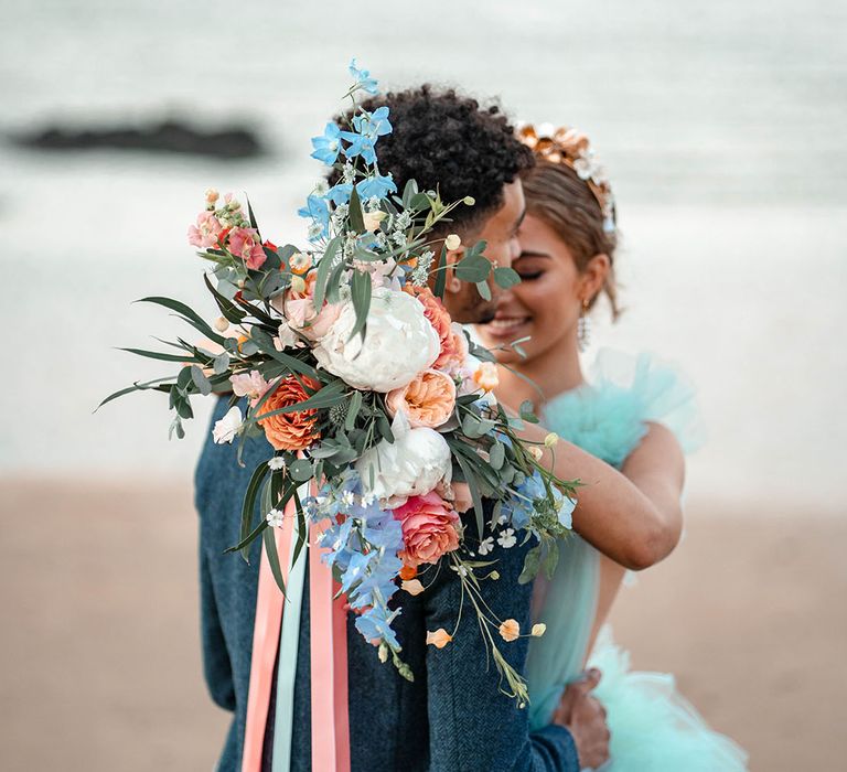 Bride holding her pastel bouquet tied with ribbon over her grooms shoulder with white peonies, coral and peach roses and blue wild flowers