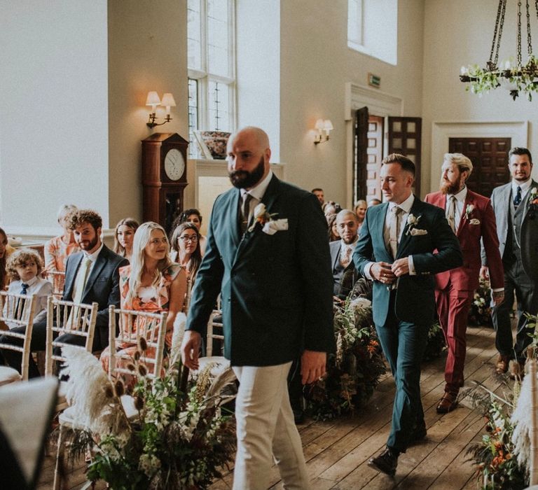 Groom walks down the aisle wearing deep green jacket and floral buttonhole