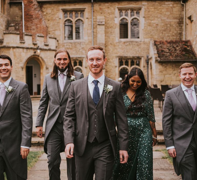 Groom in grey three piece suit with white rose buttonhole walks with three groomsmen in grey morning suits and woman in emerald sequin dress in the grounds of Notley Abbey