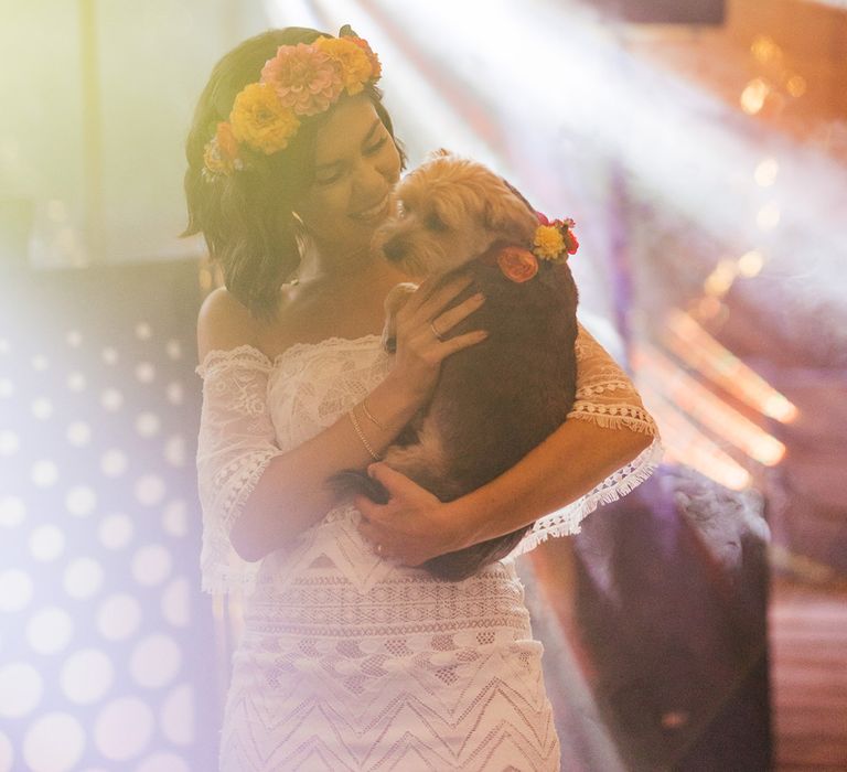 Bride in off the shoulder Grace Loves Lace wedding dress and flower crown holds dog in floral dog collar inside barn for evening wedding reception