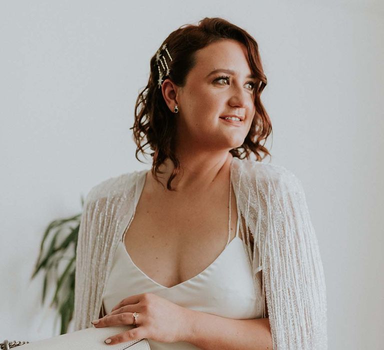 Bride wears her short hair slightly curled whilst holding white bag and wearing tassel Halfpenny London wedding cape