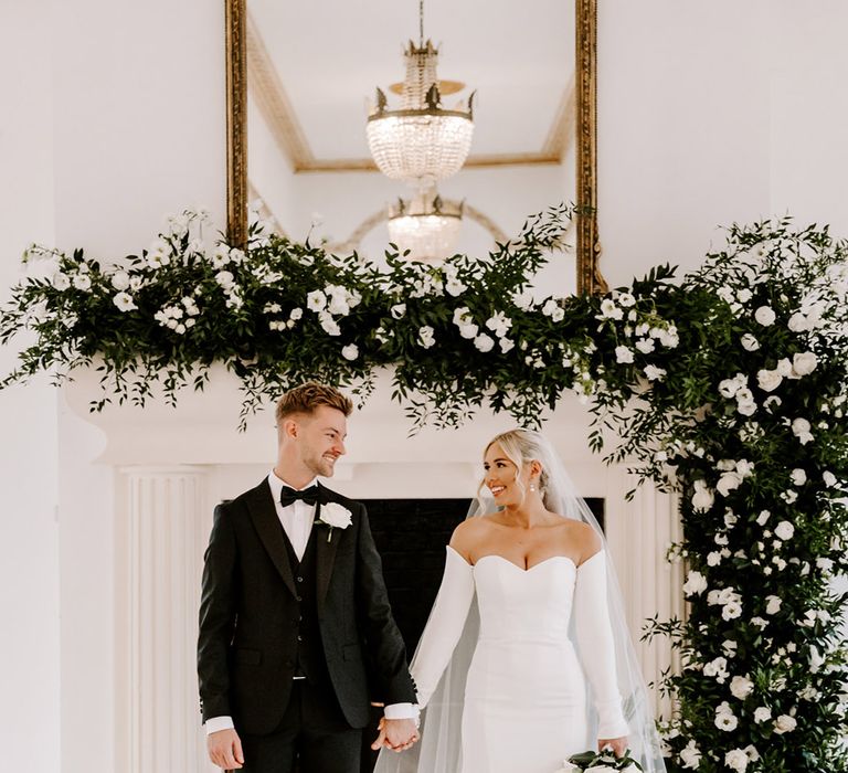 Bride in a strapless wedding dress with detachable long sleeves holding hands with her groom in a tuxedo in front of the fireplace decorated in green foliage and white flowers at Northbrook Park 