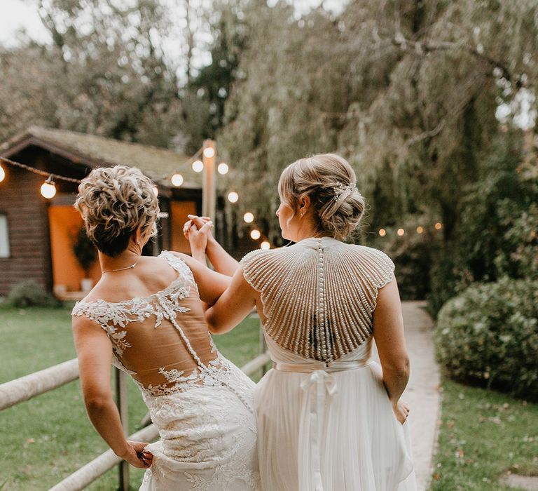 Brides hold hands as they wear bridal gowns complete with lace and beading at the back