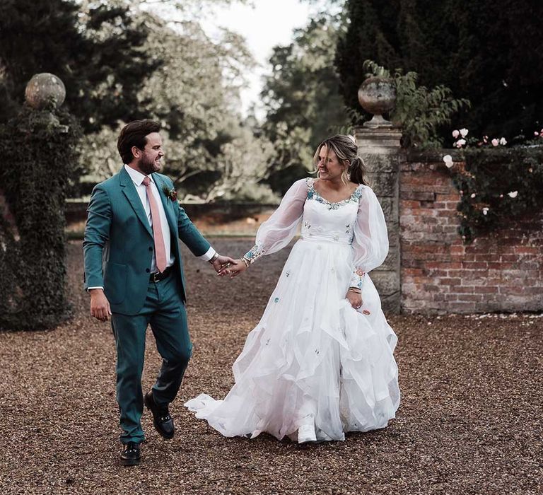 Groom in blue suit and pink tie holding hands with bride in long mesh sleeve wedding dress walks across drive at Iscoyd Park