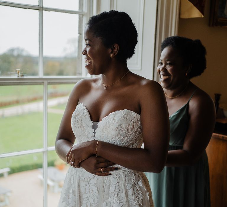 Bridesmaid in silk green dress helps beautiful bride get ready in her off the shoulder wedding dress 