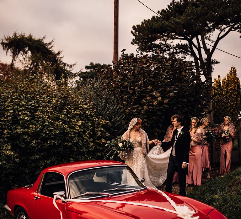 Bridesmaids help the bride to the vintage red car at Oxnead Hall wedding