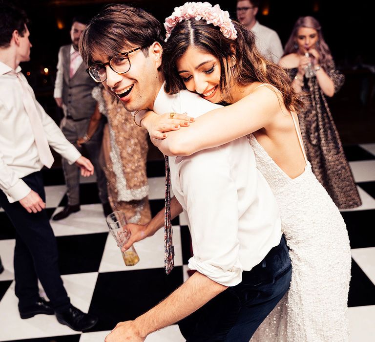 Bride in pink flower crown headband and beaded midi dress hugs groom holding beer on the dance floor for evening reception 