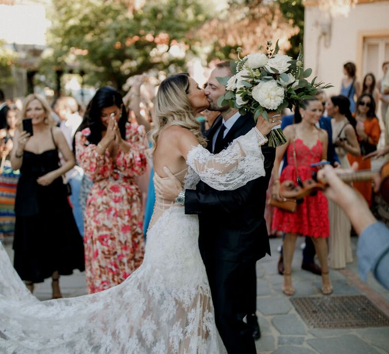 Bride & groom kiss outdoors as wedding guests watch on 