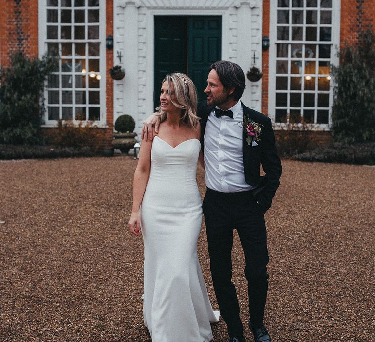 Groom wraps his arm around shoulders of his bride as they stand outdoors in front of Gosfield Hall 