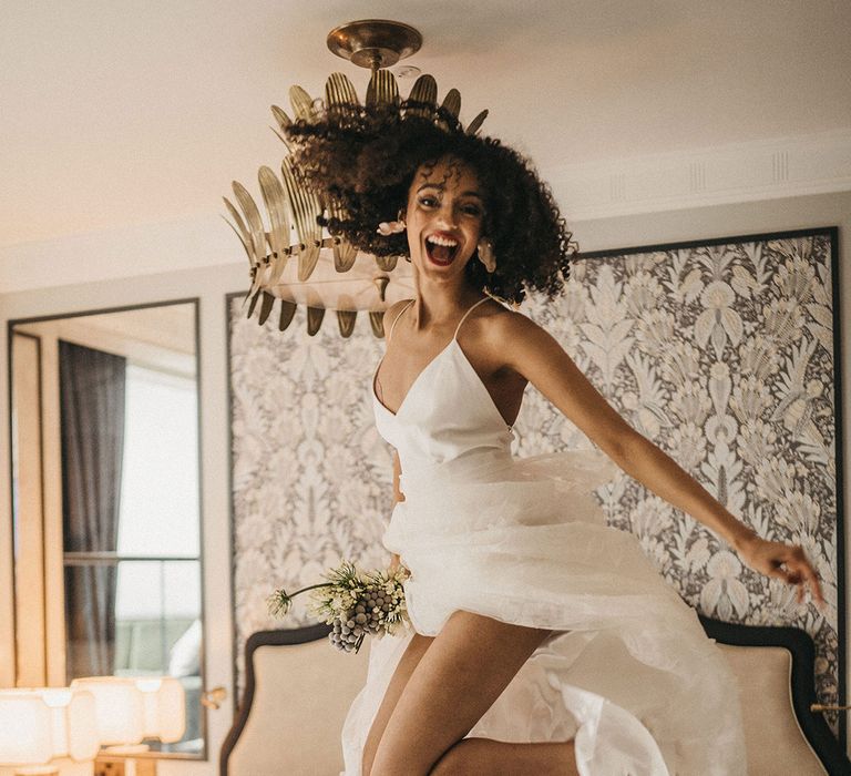 Black bride with naturally curly hair jumping on the bed at Mayfair Townhouse in a spaghetti strap wedding dress with lace skirt 