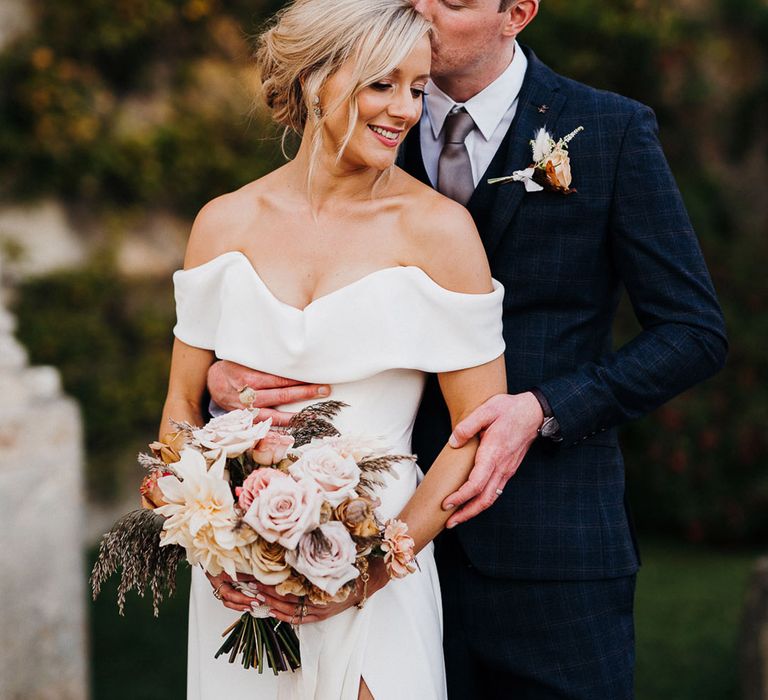 Groom in blue checkered suit hold wife and kisses her forehead with bride in sweetheart off the shoulder front slit style wedding dress 