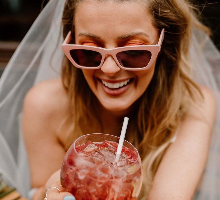 Bride in a retro pink sunglasses holding a ballon glass filled with a fruity cocktail, ice and a straw 