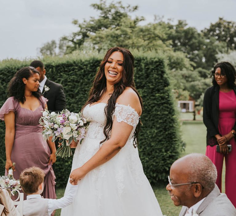 Gorgeous smiling bride in off the shoulder lace wedding dress, long lace veil and purple and white wedding bouquet 