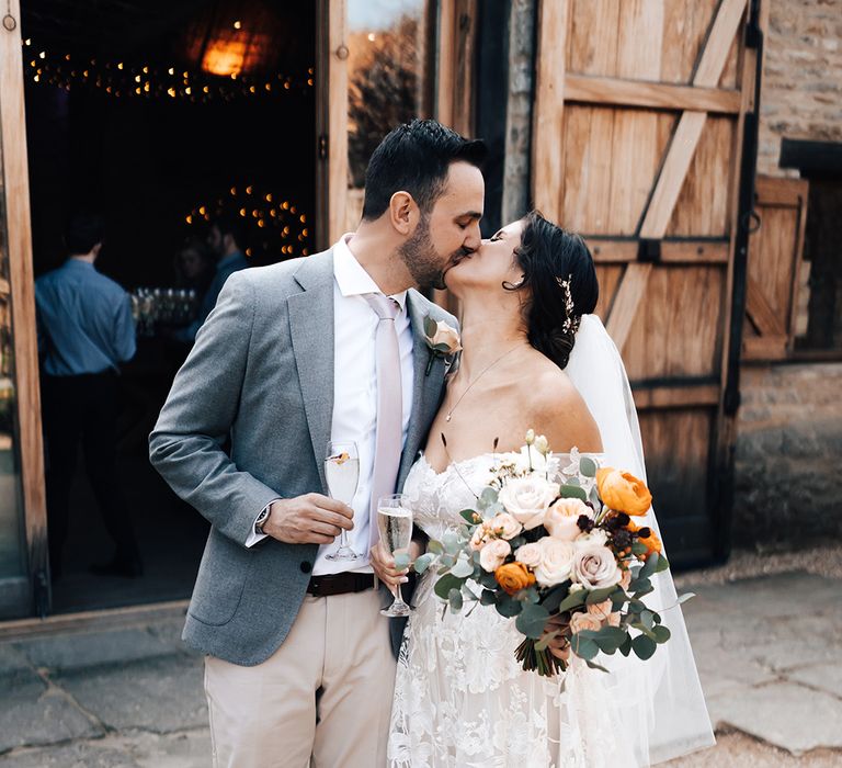 Bride and groom kiss holding glasses of champagne and a bouquet of orange and white autumnal flowers including roses and peonies 