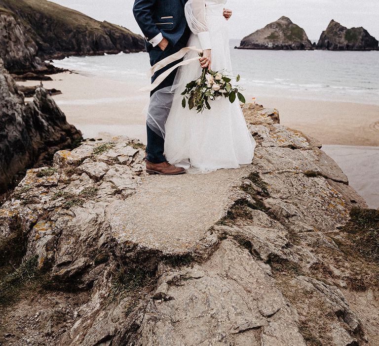 Bride and groom stand on rock with a view as he groom his the bride from behind and kisses her forehead