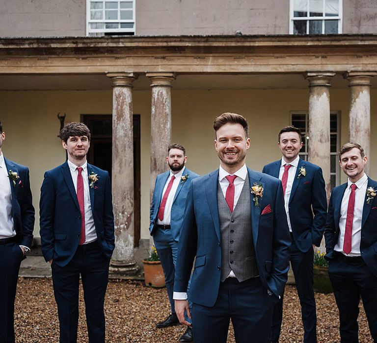 Groom in three piece grey and blue suit with red tie with groomsmen in matching navy blue suits and red ties