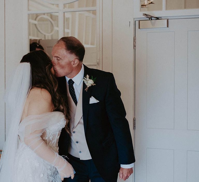 Father of the bride in dark suit and beige waistcoat kisses the bride as he sees her in her wedding dress