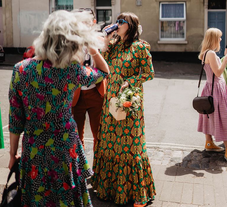 Bride wears blue heart shaped glasses and holds bridal bouquet whilst sipping beer outdoors 