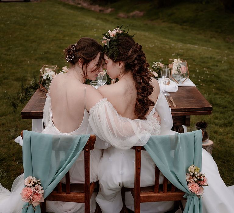 Two brides sitting at their outdoor sweetheart table decorated with drapes, pink flowers and lanterns 