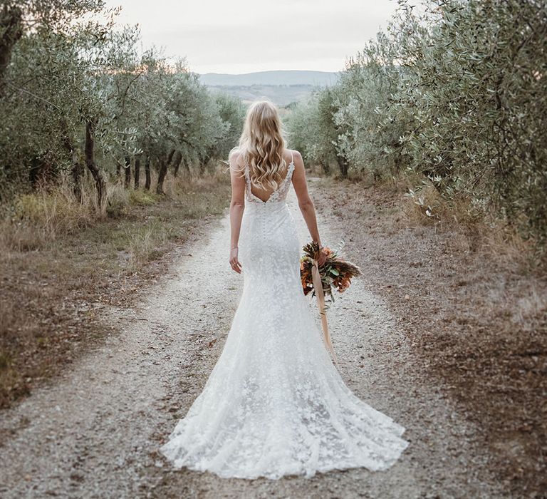 Bride walks through Tuscan countryside holding floral bouquet and wearing romantic lace wedding dress with low back