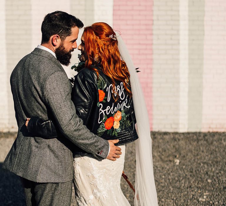 Bride in personalised leather jacket cover-up puts an arm around the groom in a grey three piece suit 