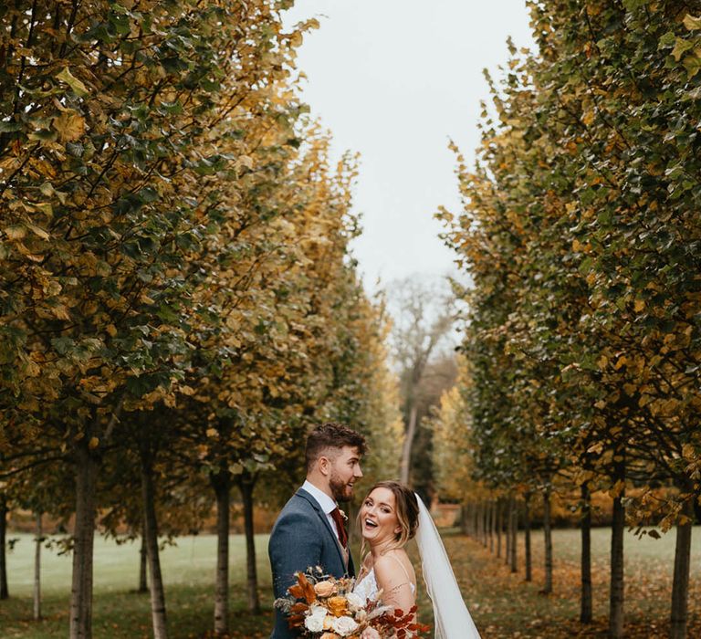 Bride holding autumnal wedding bouquet stands with the groom amongst autumn trees 