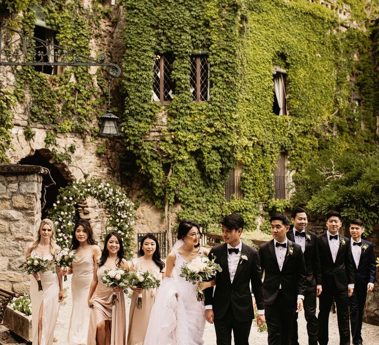 Bride & groom stand with their wedding party and bridesmaids who wear champagne coloured dresses in different styles whilst holding pastel floral bouquets