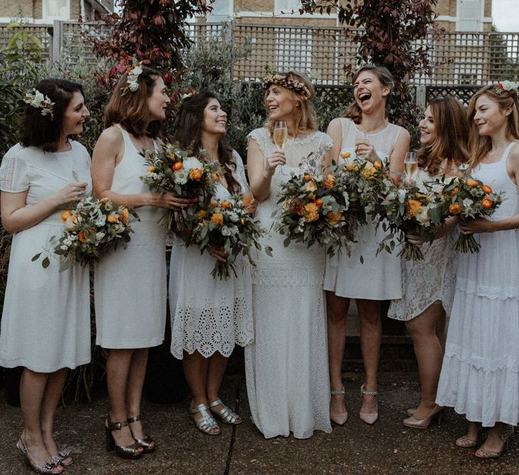 Bridal party in varying lengths of lace white bridesmaid dresses and orange and white floral bouquets 