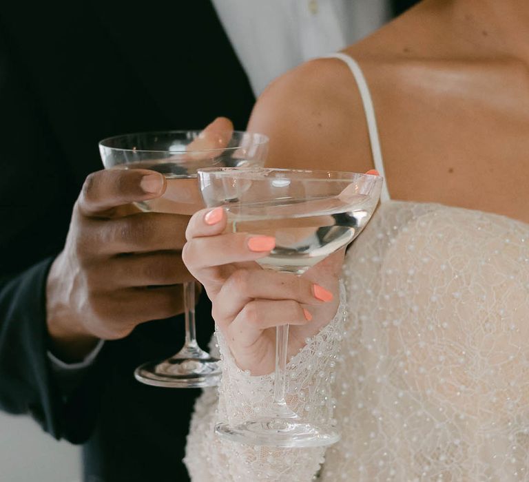 Bride with coral pink nails and groom both hold cocktail glasses as they drink together 