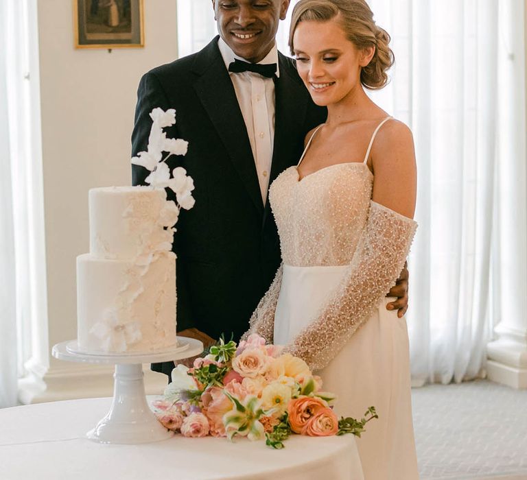 Bride in evening wedding dress with beads and off the shoulder detail with groom in black tie as they look to cut the cake 