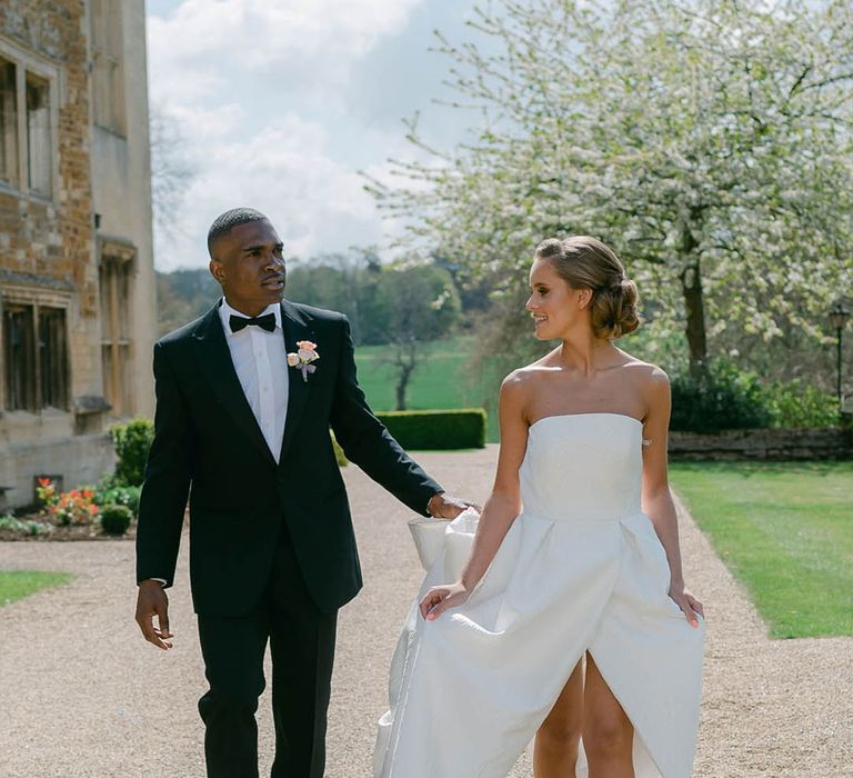 Groom in black tie walks holding the train of the bride's wedding dress as they walk together 