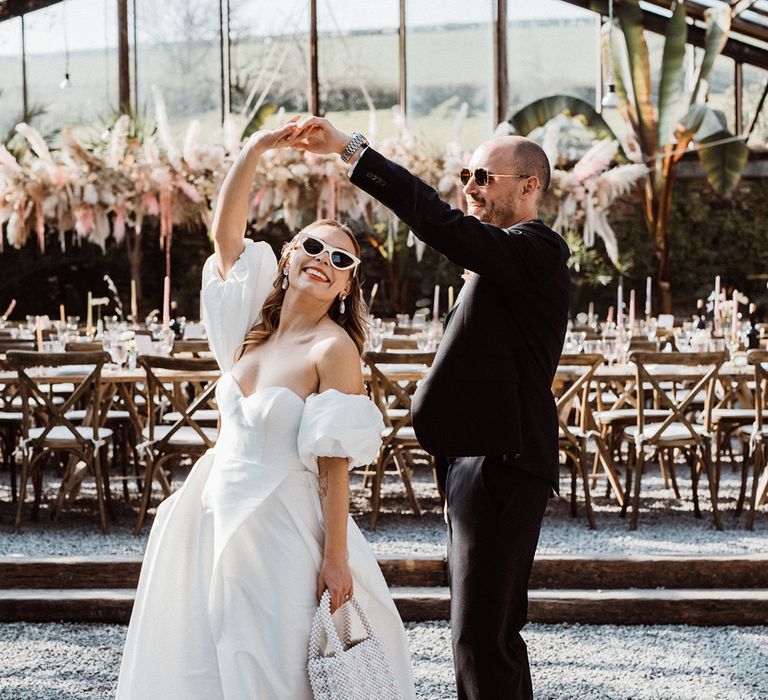 Groom in navy suit spins around the bride in a puff sleeve wedding dress with sunglasses and a pearl handbag 