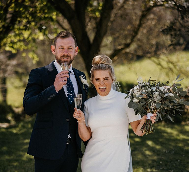 Groom in blue suit with a polka dot tie with bride in a high neck Jesus Peiro wedding dress having glasses of champagne 