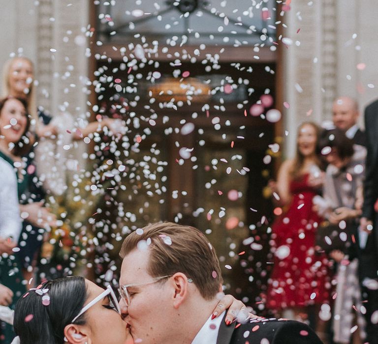 Bride wears pearl hoop earrings and holds bright floral bouquet as she kisses her groom as colourful confetti falls around them