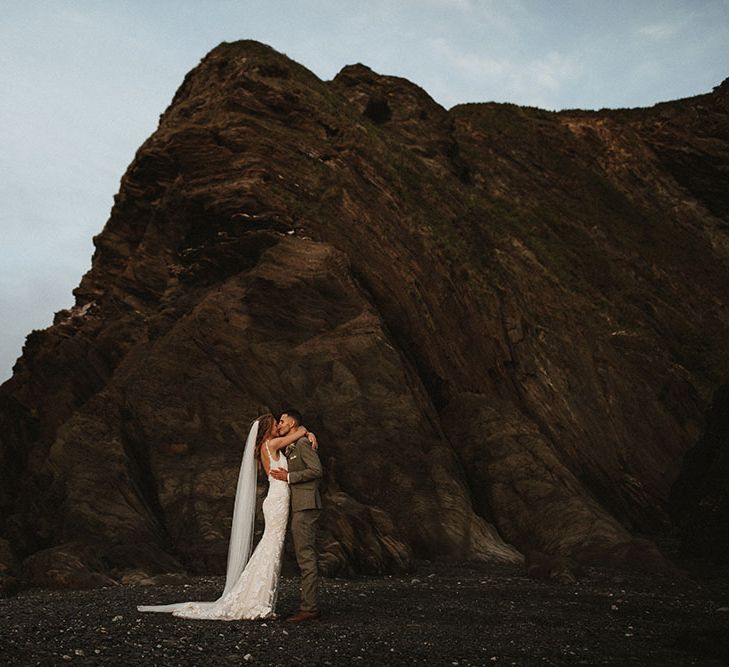 Bride & groom kiss at Tunnels Beach in Devon surrounded by dark rocks after outdoor wedding ceremony