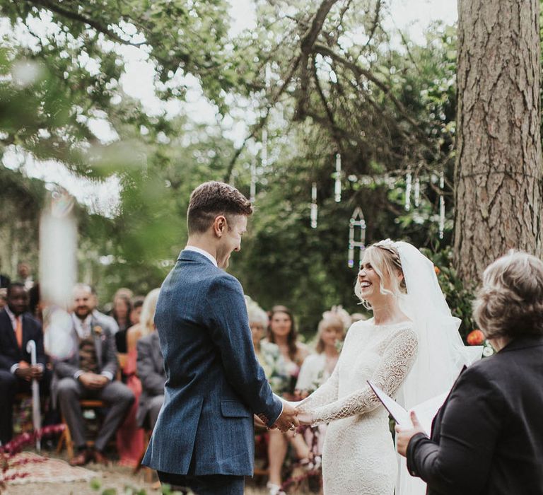 Bride in a lace long sleeve wedding dress with the groom in a blue suit holding hands for their outdoor woodland ceremony 