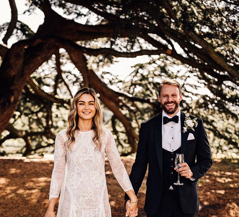 Groom in black tie holding champagne and the bride's hand with the bride in a long sleeve lace boho wedding dress with pet dog 