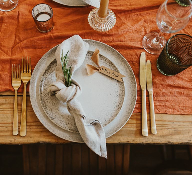 Place setting with gold cutlery, white napkins, orange table runner, and speckled plates 