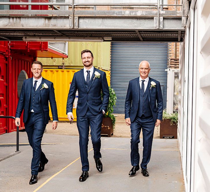 Groom wears three-piece blue suit complete with white floral buttonhole as he walks with his groomsmen in matching suits 