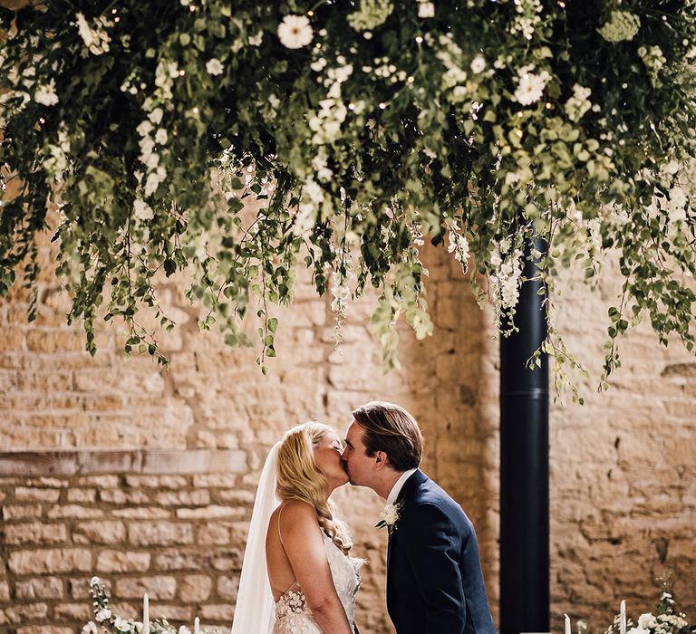 Bride in an Alexandra Grecco Lana dress and groom in a navy blue suit share their first kiss as a married couple 