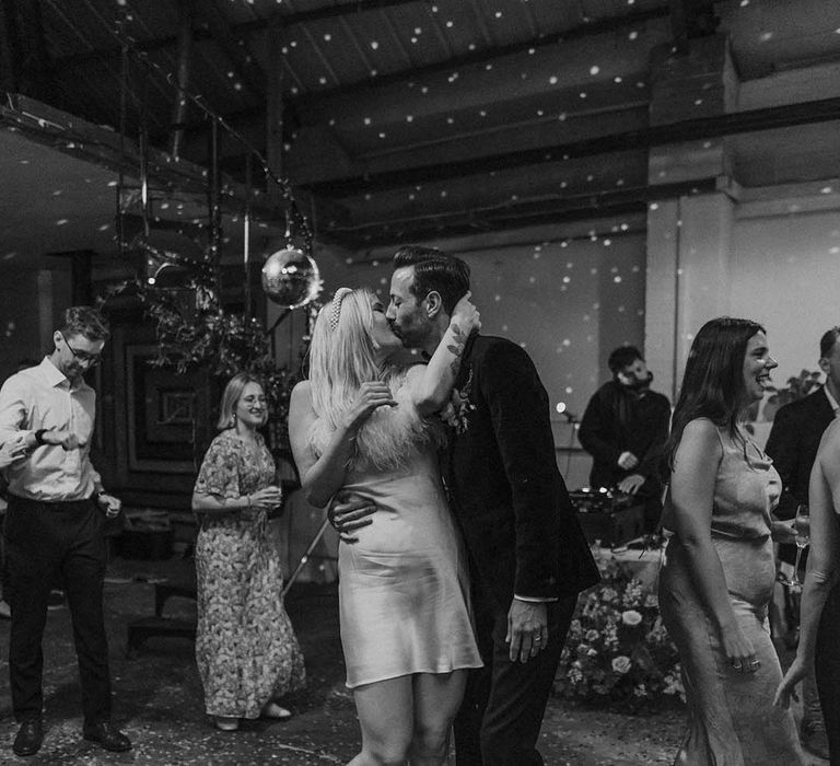 Bride in short satin dress with feather trimmed neckline and groom in dark tux kissing at Hackney Studios